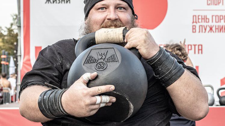 a man holding an 88kg kettlebell and biting on the leather strap over the handle.