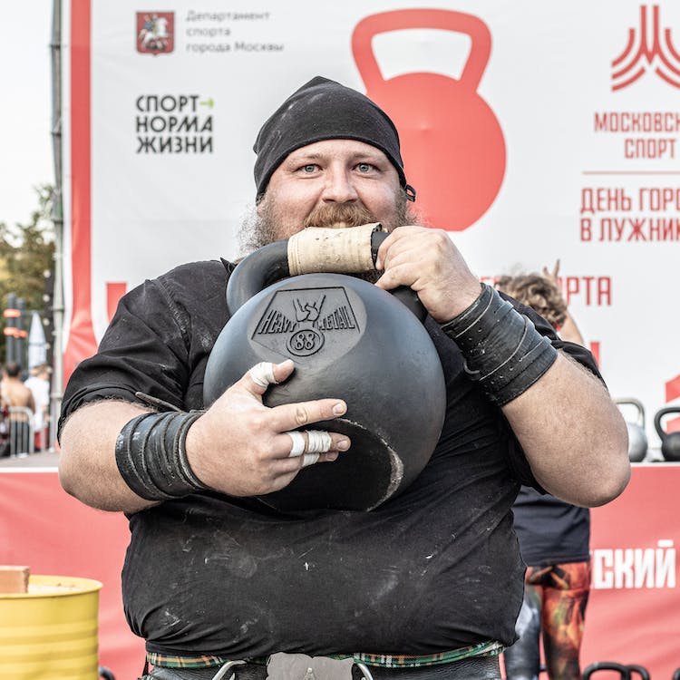 a man holding a 88kg kettlebell in his hands and biting the leather strip over the handle